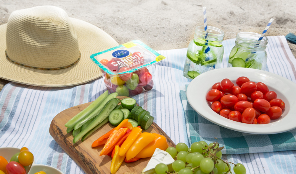 Family beach picnic. A blanket on the sand with a sun hat, cucumber water with paper straws in mason jars, and a range of fruits and vegetables for snacking including grape tomatoes, mini cucumbers, and mini sweet peppers.