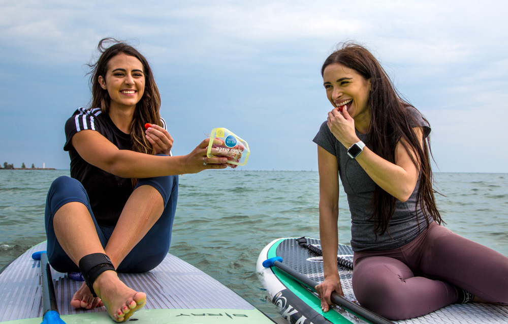 Two girls sitting side by side on paddle boards in the open water sharing a package of red grape tomatoes as a snack.